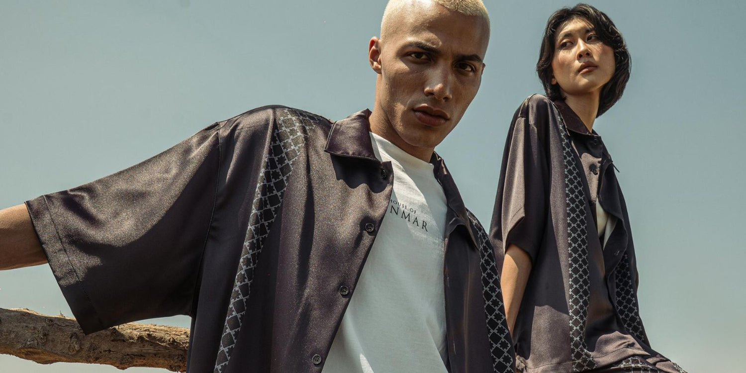 Two models wearing matching black short-sleeve button-up shirts with geometric patterned panels from House of Cenmar, styled in a modern streetwear look under a clear sky.