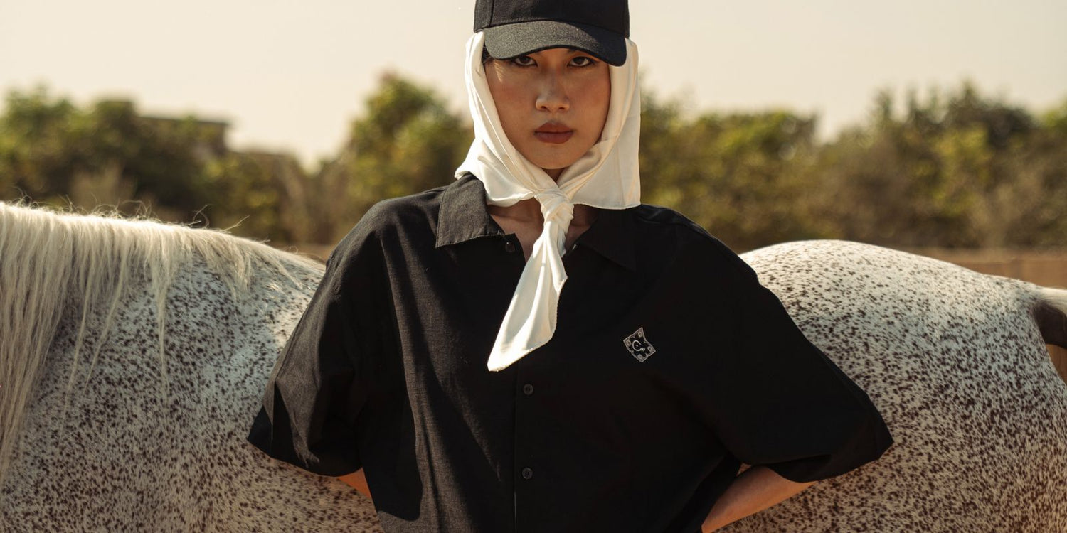 Female model wearing a black short-sleeve button-up shirt from House of Cenmar, styled with a black cap and white scarf, standing beside a spotted horse.