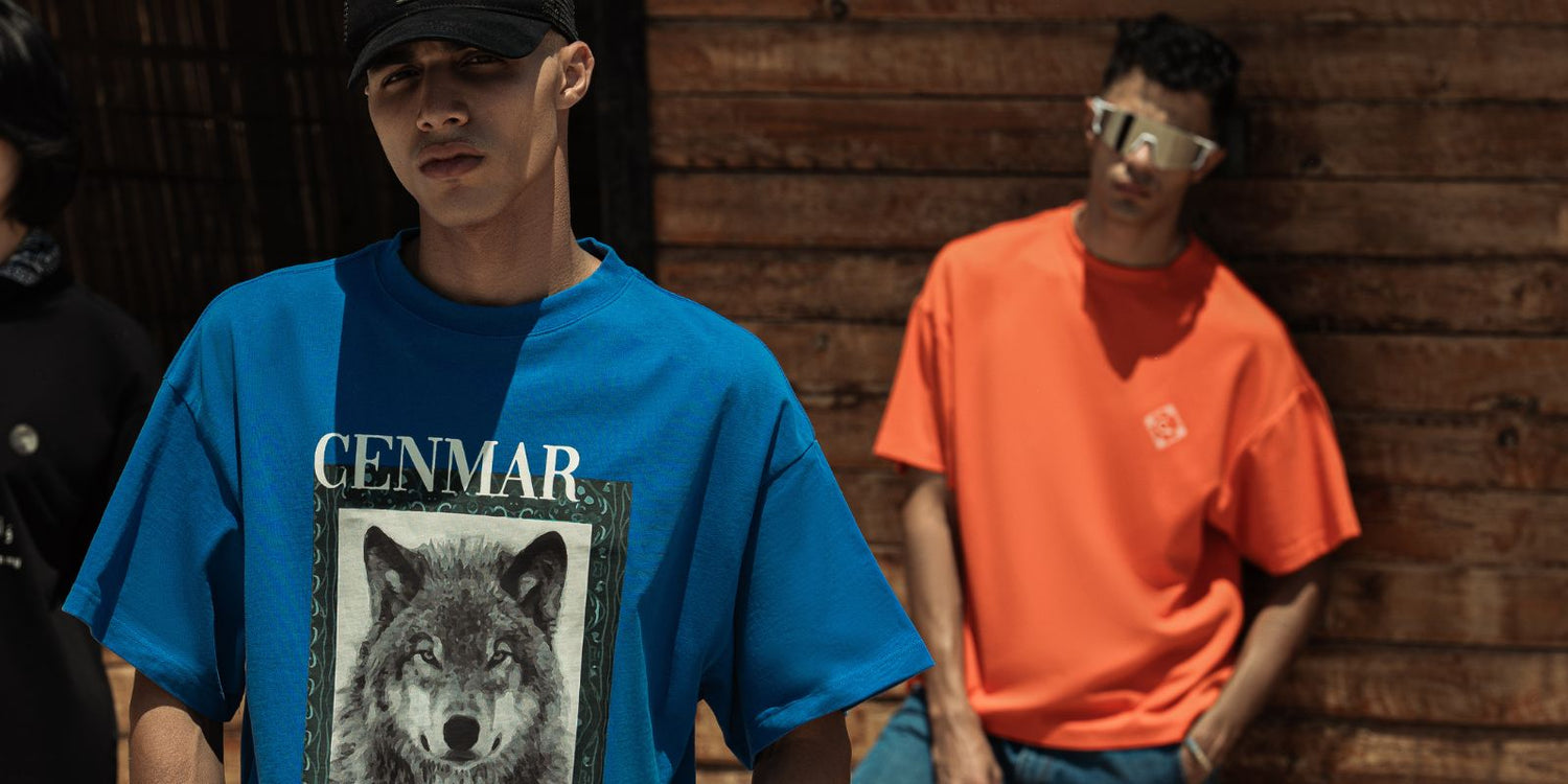 Two male models in oversized streetwear standing against a wooden wall. The model in front wears a blue graphic wolf t-shirt with “CENMAR” text and a black cap, while the model behind wears an orange oversized t-shirt, jeans, and reflective sunglasses.